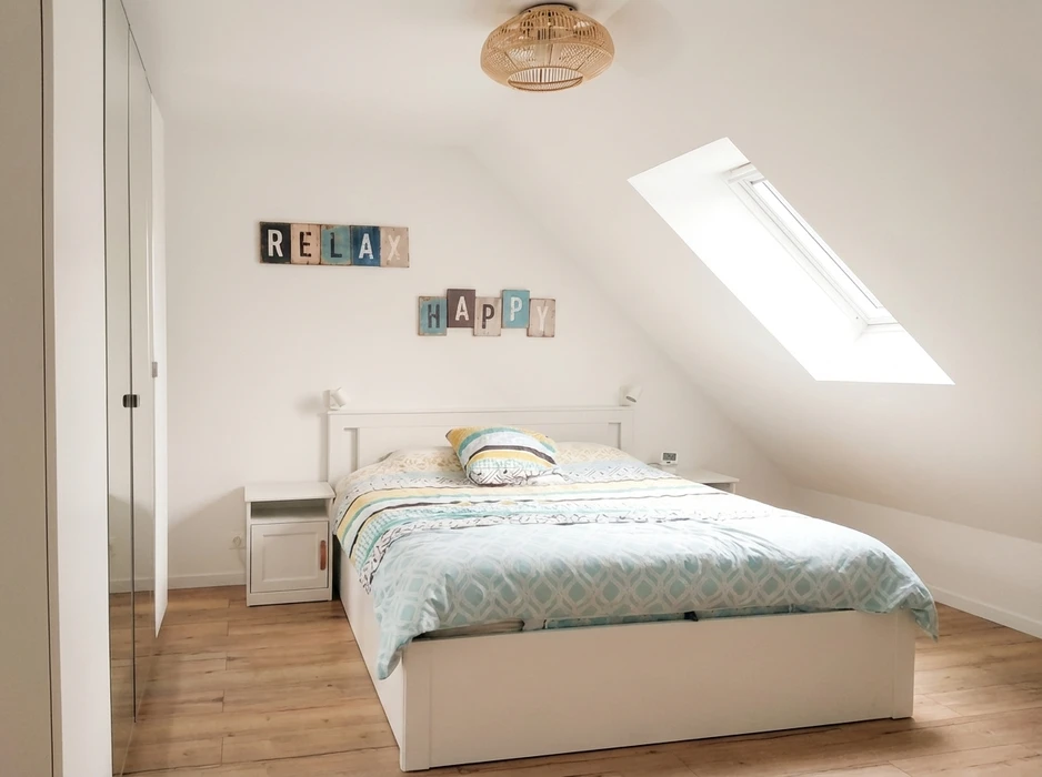 Chambre mansardée lumineuse avec lit double blanc, parure de lit géométrique bleu et jaune, deux tables de chevet blanches