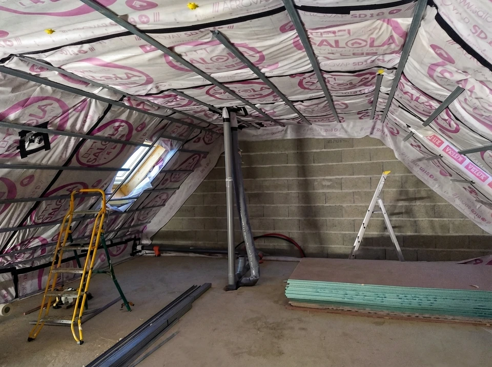 Ouvrier en combinaison orange et casque rouge installe une membrane d'étanchéité noire sur charpente en construction